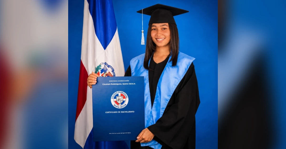 Graduate in a black cap and blue gown holding a blue diploma against a blue backdrop with a flag beside her person.