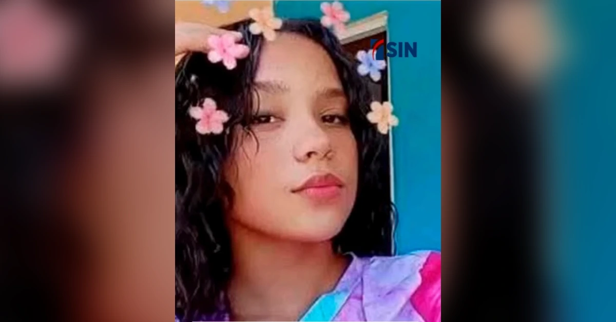 Young person with dark curly hair wearing a pink flower crown, posing for a selfie with a blue background.