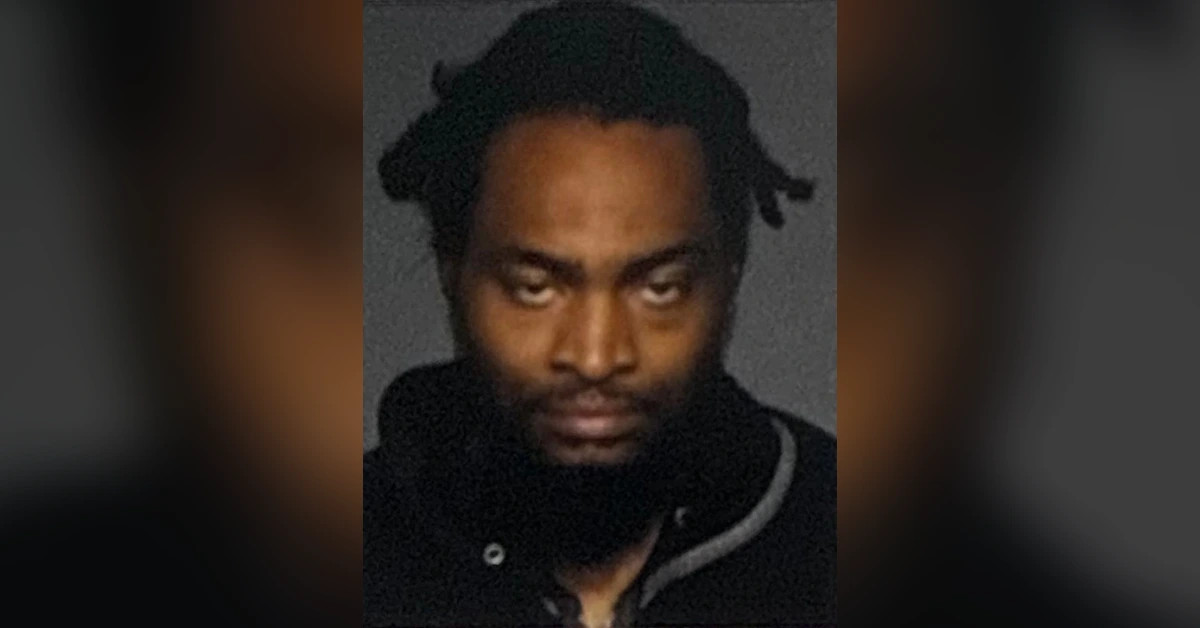 Mugshot-style photo of a man with dreadlocks, wearing a dark jacket against a gray background, looking at the camera with a stern expression.