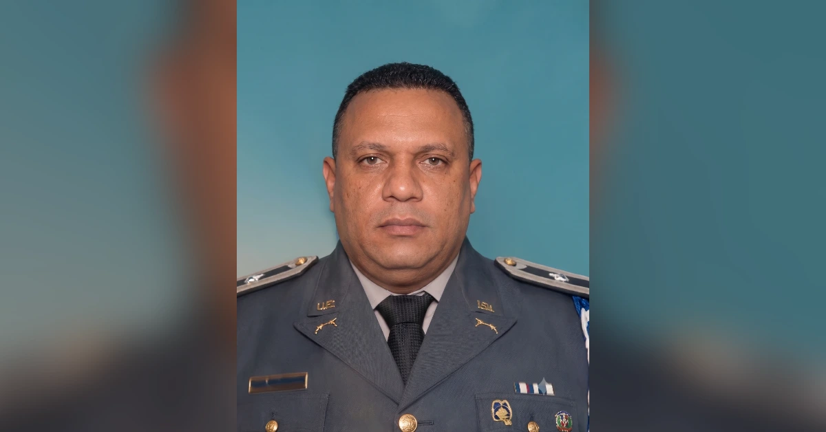 Portrait of a man in a gray military dress uniform with badges and ribbons against a teal backdrop.