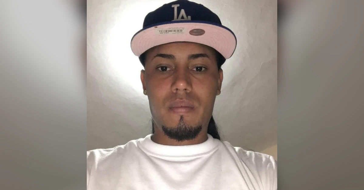 Portrait of a man facing the camera, wearing a black LA cap with a pink brim and a white T-shirt, neutral expression.