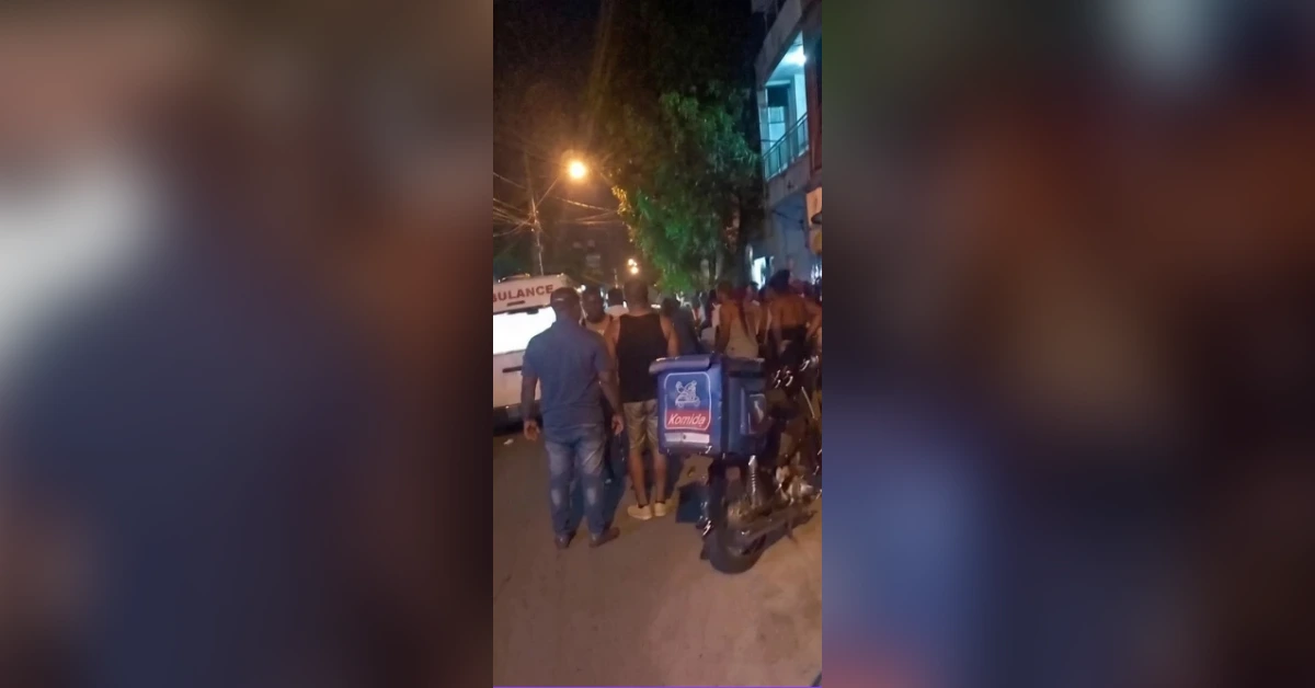 Nighttime street scene with a crowd gathered around a delivery scooter and an ambulance in the background.