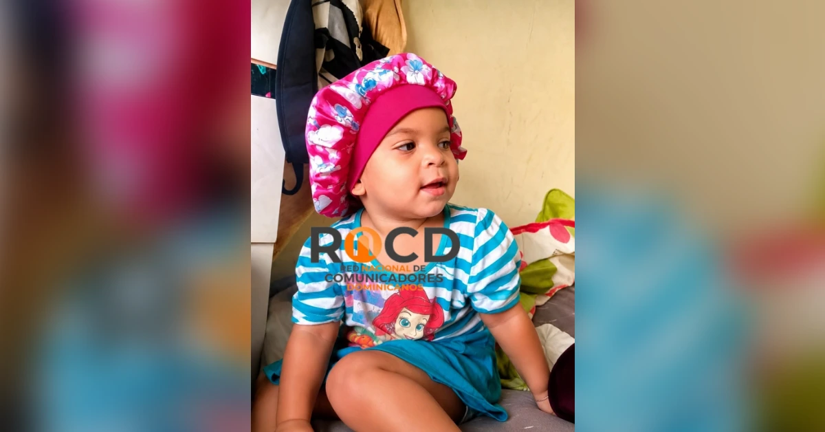 Young child wearing a pink floral bonnet and blue striped shirt, seated indoors and looking to the right.