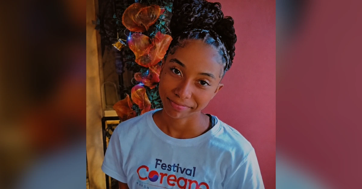 Young woman with a braided bun, wearing a light blue T-shirt, smiling, with colorful flowers in the background outdoors.
