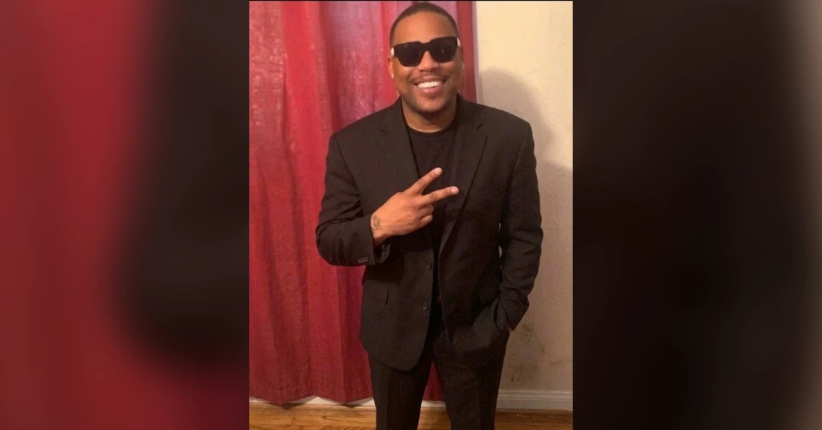 Man in a black suit and sunglasses, smiling and flashing a peace sign indoors beside a red curtain.