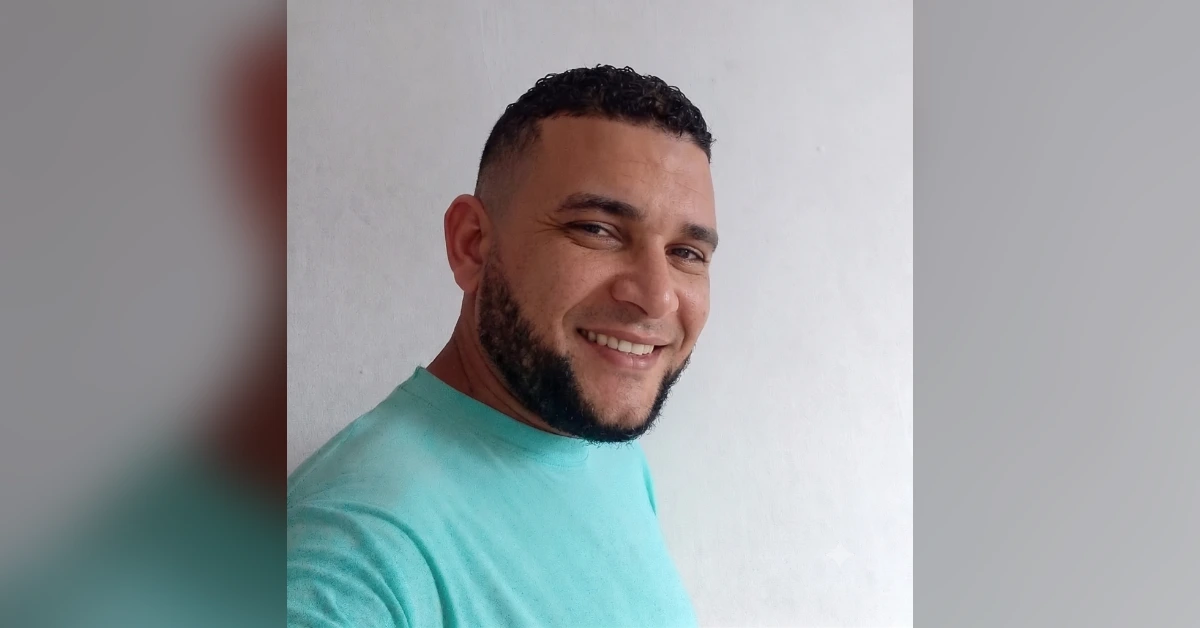 Smiling man with short dark hair and a beard in a turquoise shirt, standing against a light wall.