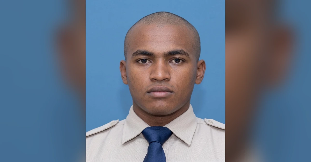 Portrait of a young man in a beige uniform shirt and navy tie against a blue background, facing the camera.