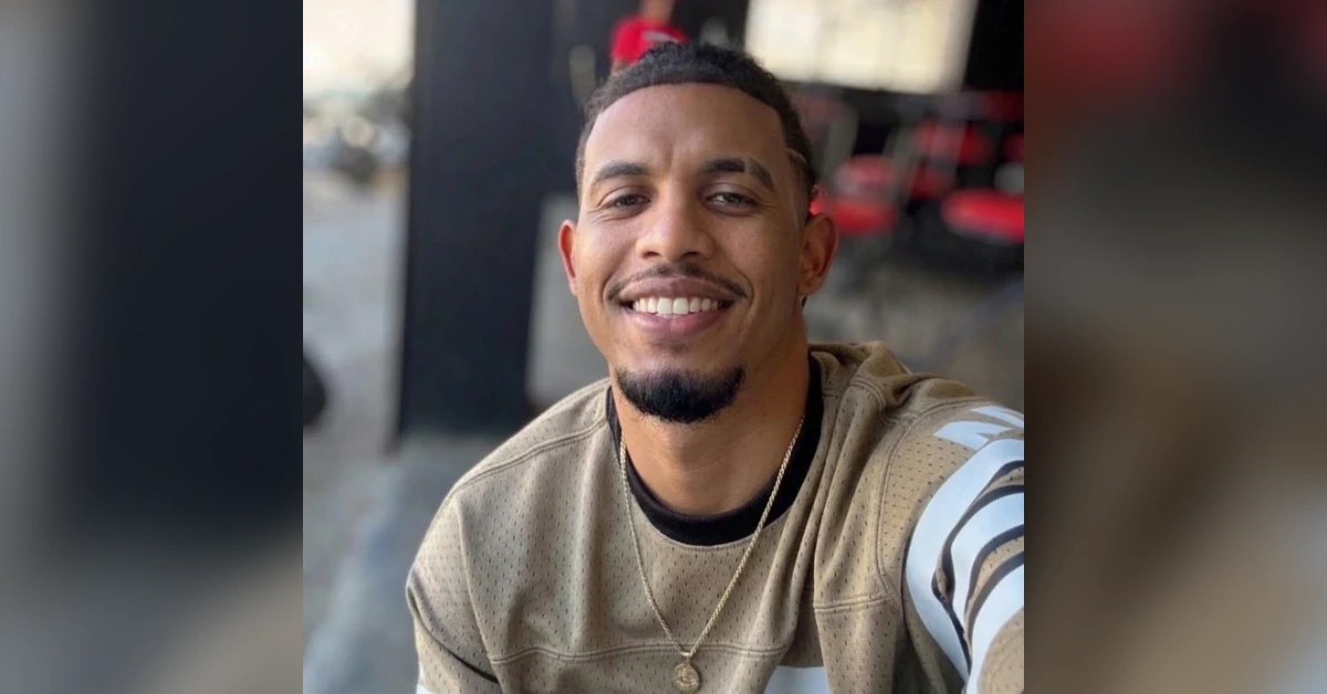 Smiling man taking a selfie, wearing a beige jersey and gold chain, in an outdoor/café setting