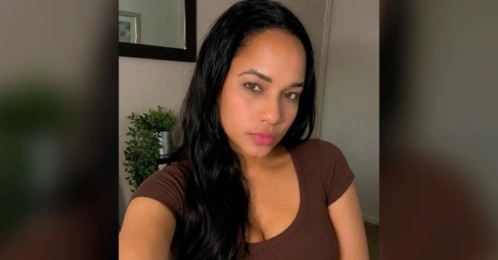 Close-up selfie of a woman with long dark hair wearing a brown V-neck top, looking at the camera.
