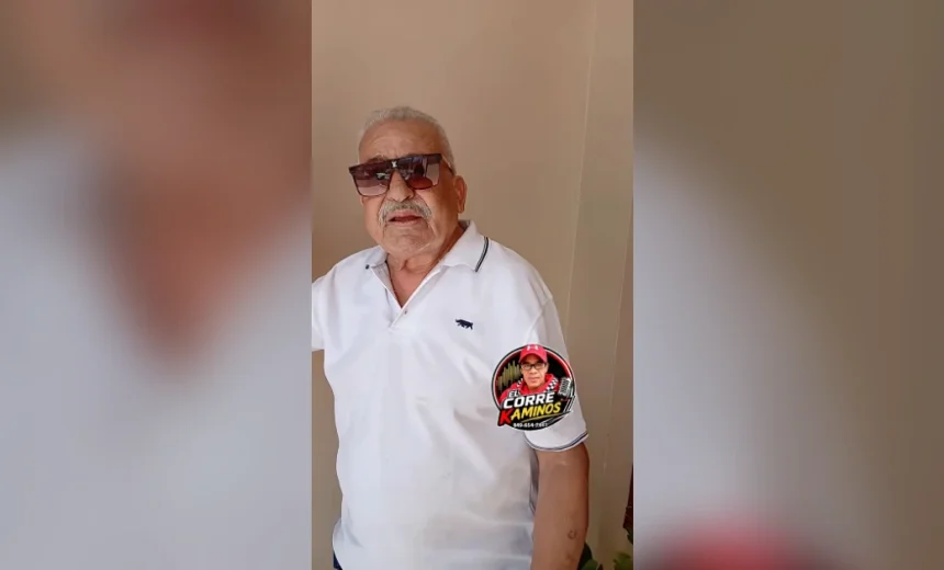Older man wearing a white polo shirt and dark sunglasses, standing indoors with neutral walls behind him; round sticker on his sleeve shows a logo.