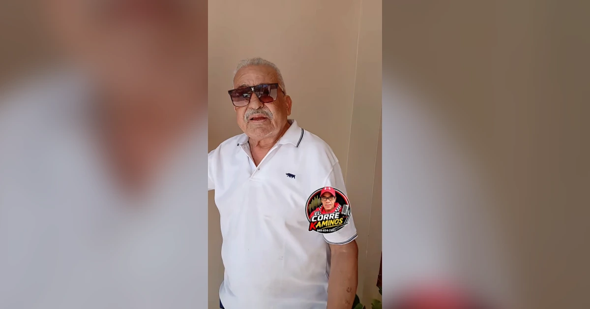Older man with gray hair and mustache wearing a white polo shirt and dark sunglasses, standing indoors.