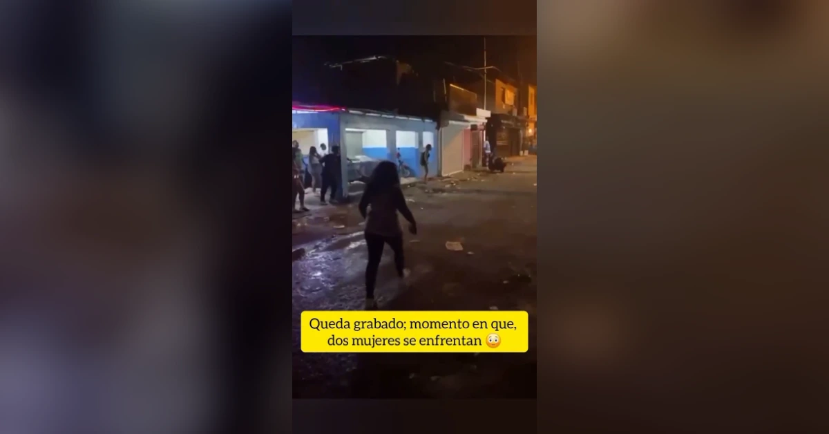 Night street scene at night; a woman walks toward a crowd outside a storefront while a confrontation between two women is captioned on screen in Spanish.