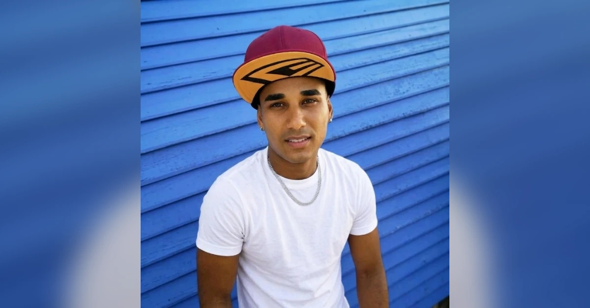 Young man wearing a white t-shirt and a red and yellow cap stands in front of a blue corrugated wall.