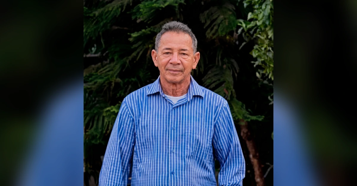 Older man in a blue striped button-down shirt standing outdoors with green foliage in the background.
