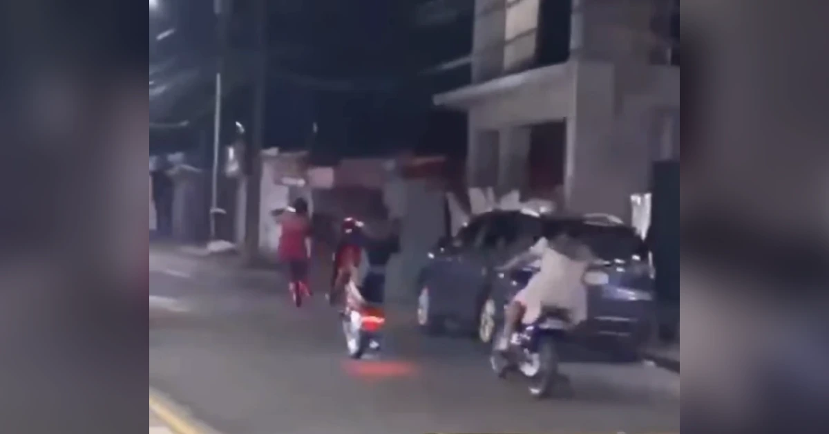 Blurry nighttime street scene with motorcyclists and pedestrians near parked cars; a person in red walks away on the left.