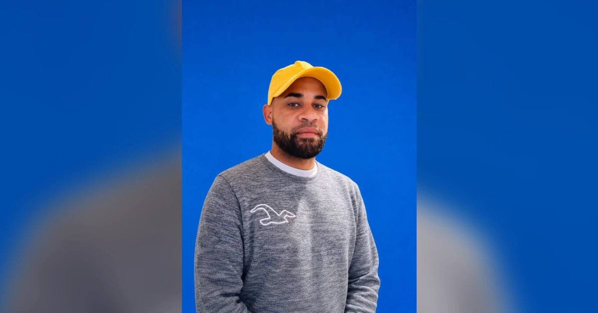 Man wearing a yellow cap and gray sweatshirt posing against a solid blue background with a neutral expression.