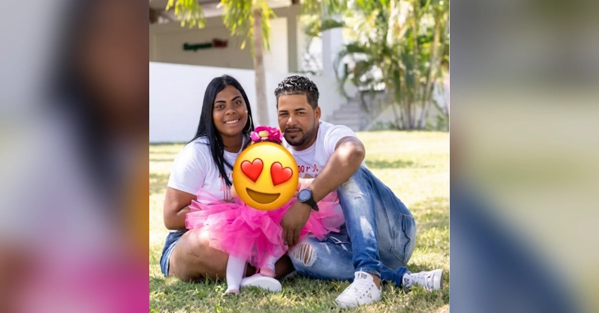 Couple sitting on the grass outdoors with their child in a pink tutu between them; emoji covers the child's face.