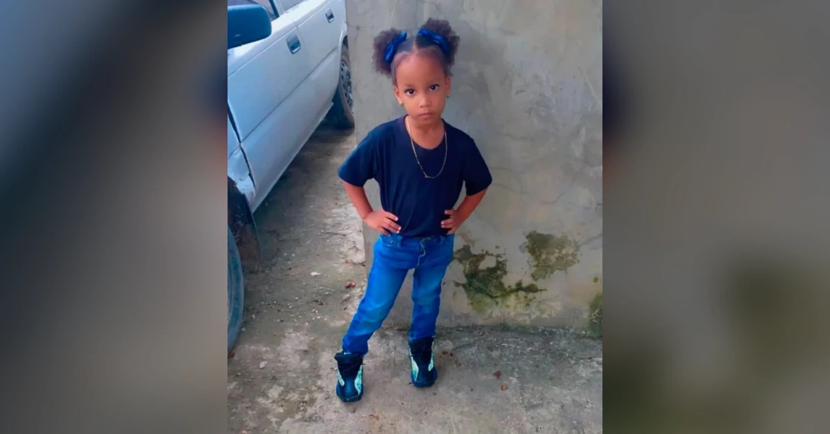 Young girl with pigtails, wearing a navy shirt and blue jeans, standing with hands on hips beside a wall.