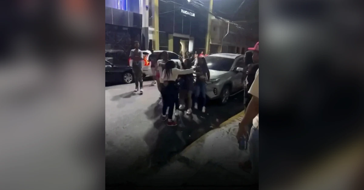 Group of people hugging in a parking lot at night, with cars parked nearby and a club entrance in the background.