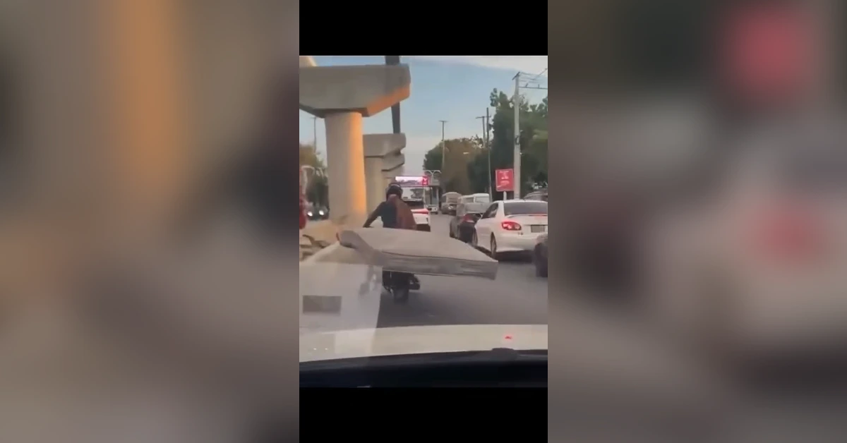 Motorcyclist balancing a large mattress on a busy street among cars, viewed from a car window.