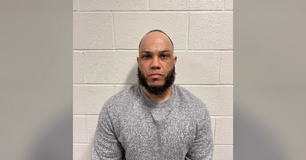 Man with a beard and shaved head facing the camera in a gray sweater against a white cinder-block wall.