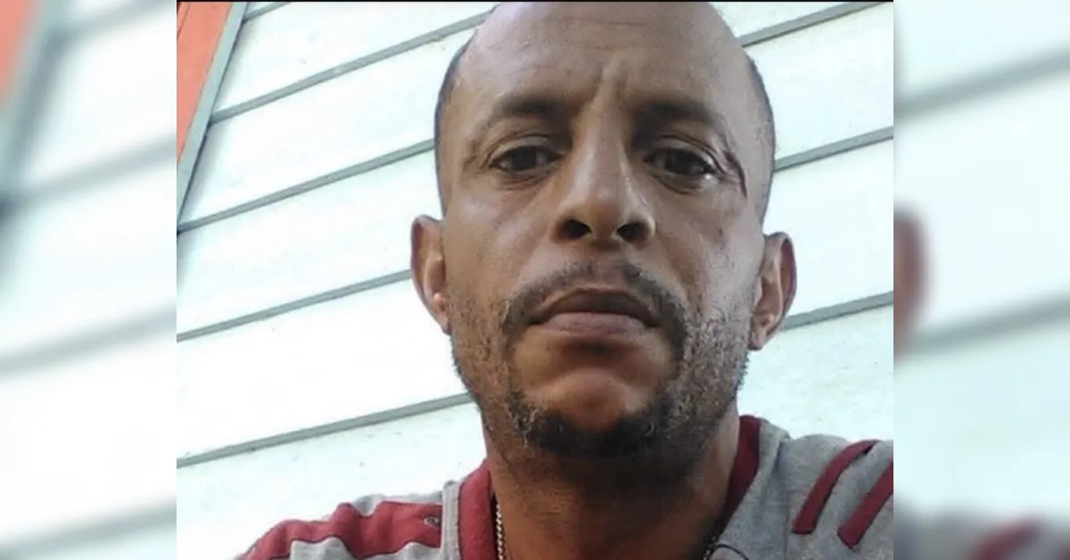 Close-up of a man with short facial hair and a striped gray-red shirt, looking at the camera against a pale siding background.