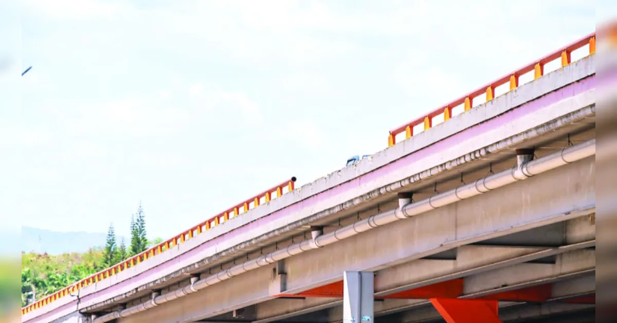 Low-angle view of an elevated concrete highway overpass with orange railing and exposed pipes, blue sky behind.