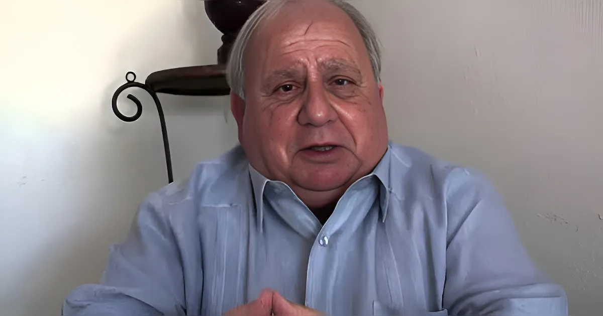Older man in a light blue shirt sitting indoors, speaking to the camera.