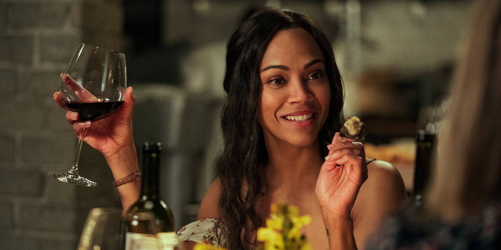 Smiling woman raising a glass of red wine in one hand and nibbling a bite of food in the other at a dinner table.