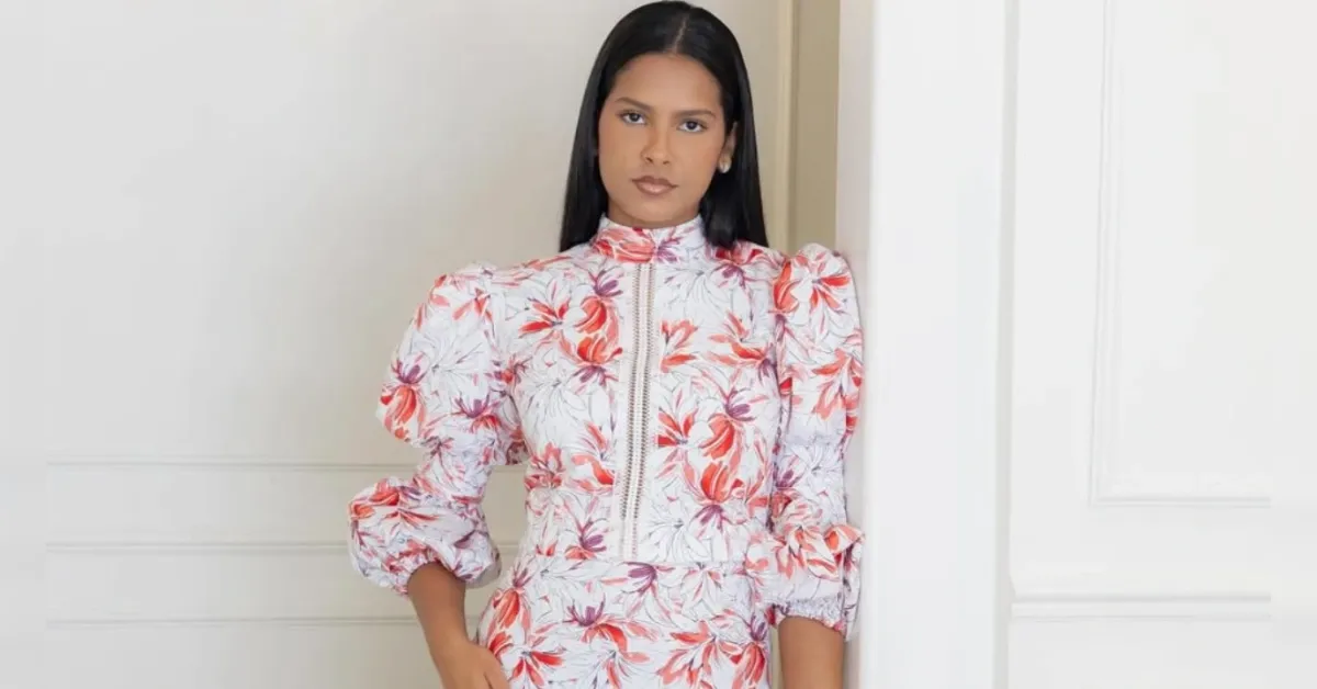 Portrait of a woman with long black hair wearing a white floral dress with puff sleeves, standing against a white wall.