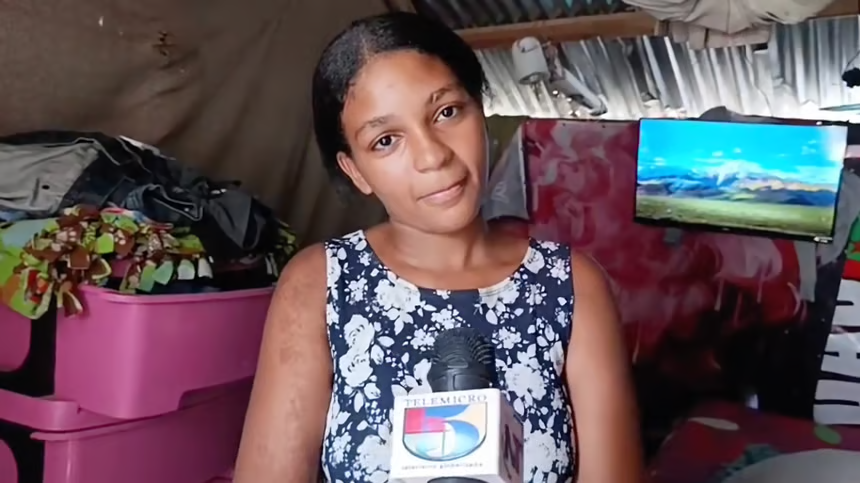 Woman in a blue floral dress being interviewed, holding a microphone with a news logo, in a cluttered room with a pink storage bin and a monitor in the background.