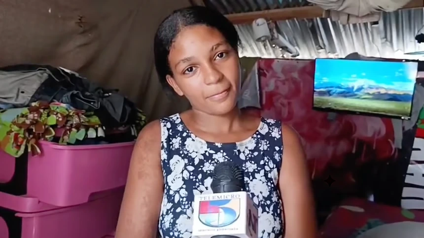 Woman in a floral sleeveless dress being interviewed indoors, holding a microphone labeled with a TV channel, with a computer monitor and cluttered background behind her.