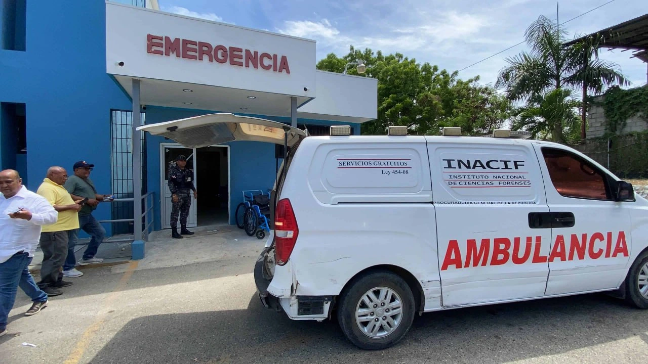 Blue hospital façade with 'EMERGENCIA' sign, as a white ambulance labeled 'AMBULANCIA' is parked at the curb and people stand nearby outside the entrance.