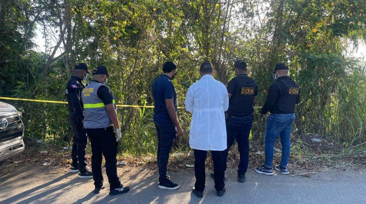 Group of investigators and police officers stand along a road behind yellow caution tape, examining a brushy area.