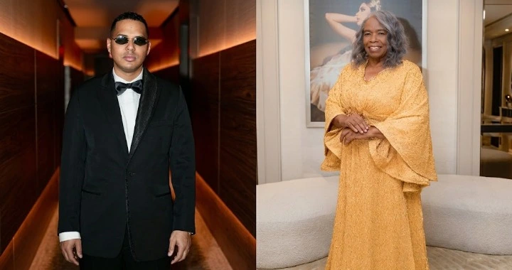 Man in a black tuxedo and sunglasses stands in a hallway beside a woman in a gold lace gown posing on a beige sofa area, indoors at a formal event venue.