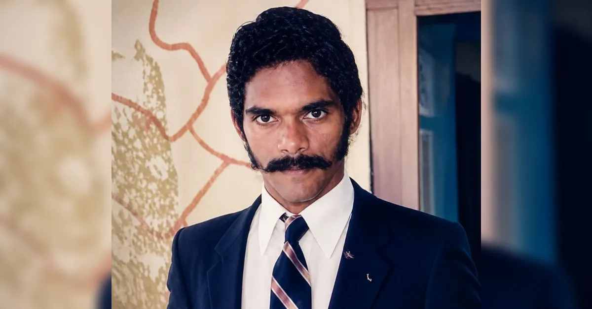 Portrait of a man with a thick mustache wearing a navy suit and striped tie, standing indoors.