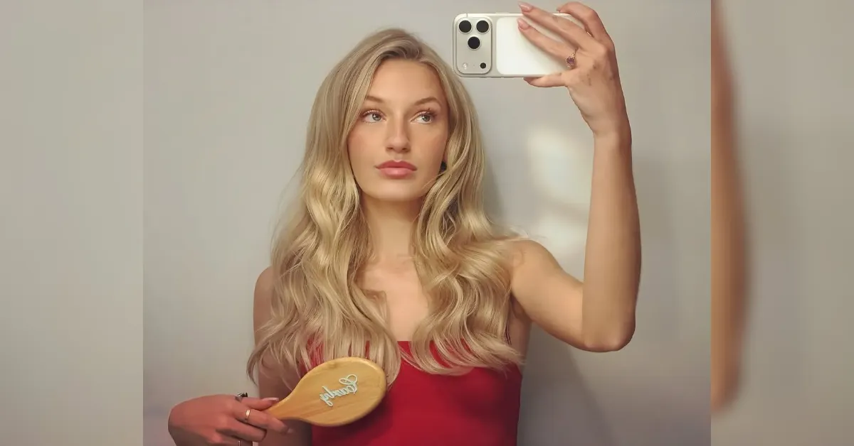 Self-portrait of a blonde woman in a red top taking a mirror selfie, holding a wooden hairbrush with wavy hair.