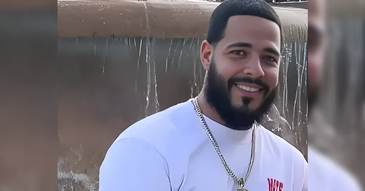Smiling man with a beard in a white shirt and gold chain, outdoors near a cascading fountain.