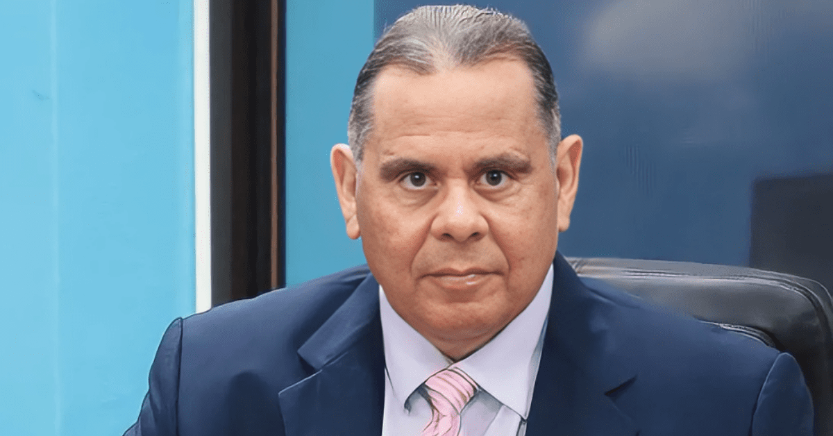 Professional head-and-shoulders portrait of a middle-aged man in a navy suit and pink striped tie, seated in an office setting with a blue background.