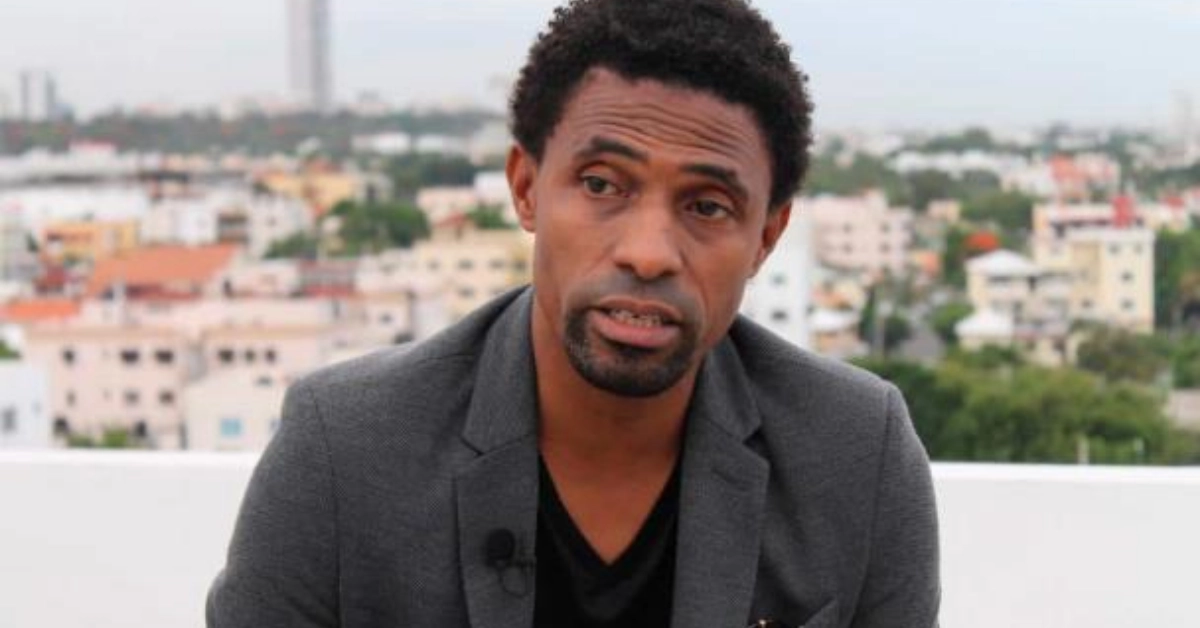 Man in a gray blazer speaks on a rooftop with a blurred city skyline behind him.