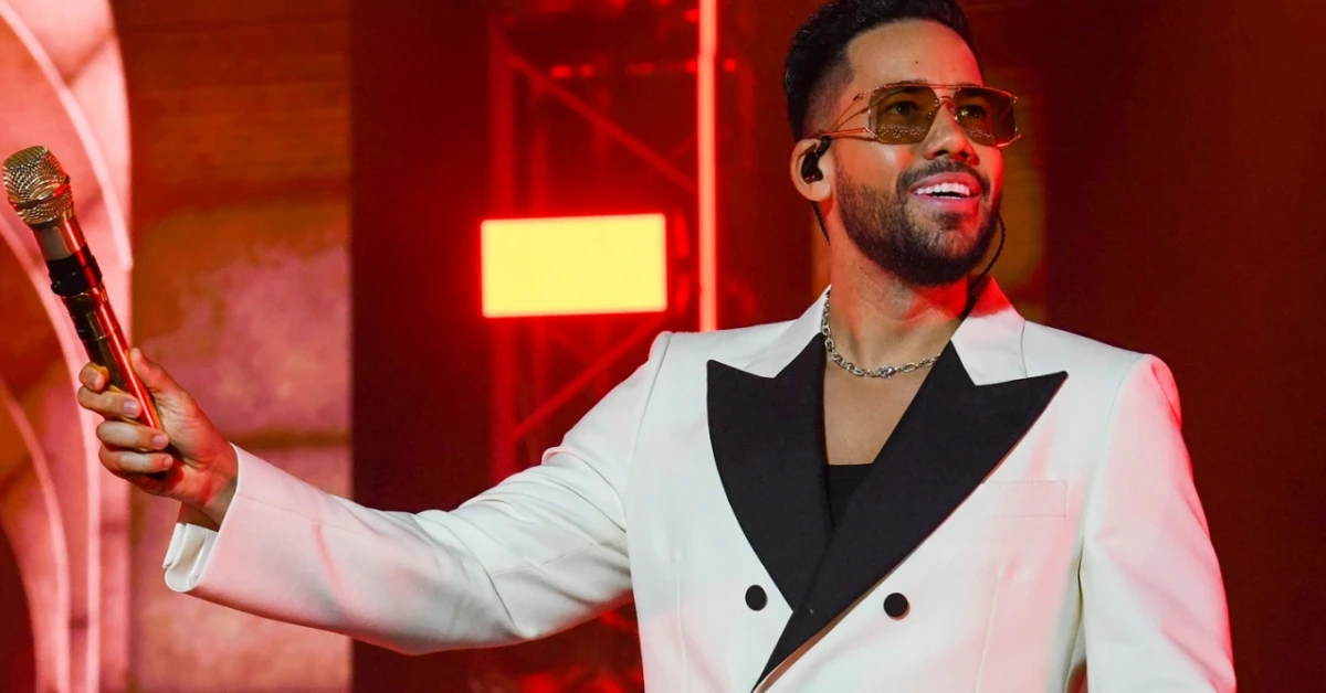 Male singer in a white suit with black lapels performs on stage, holding a microphone, wearing sunglasses and an in-ear monitor under red lights.