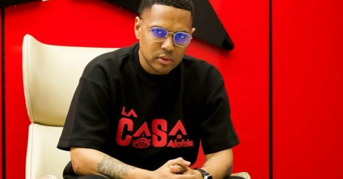 Man wearing a black T-shirt with red logo sits in a white chair against a red wall, hands clasped, wearing blue-tinted glasses.
