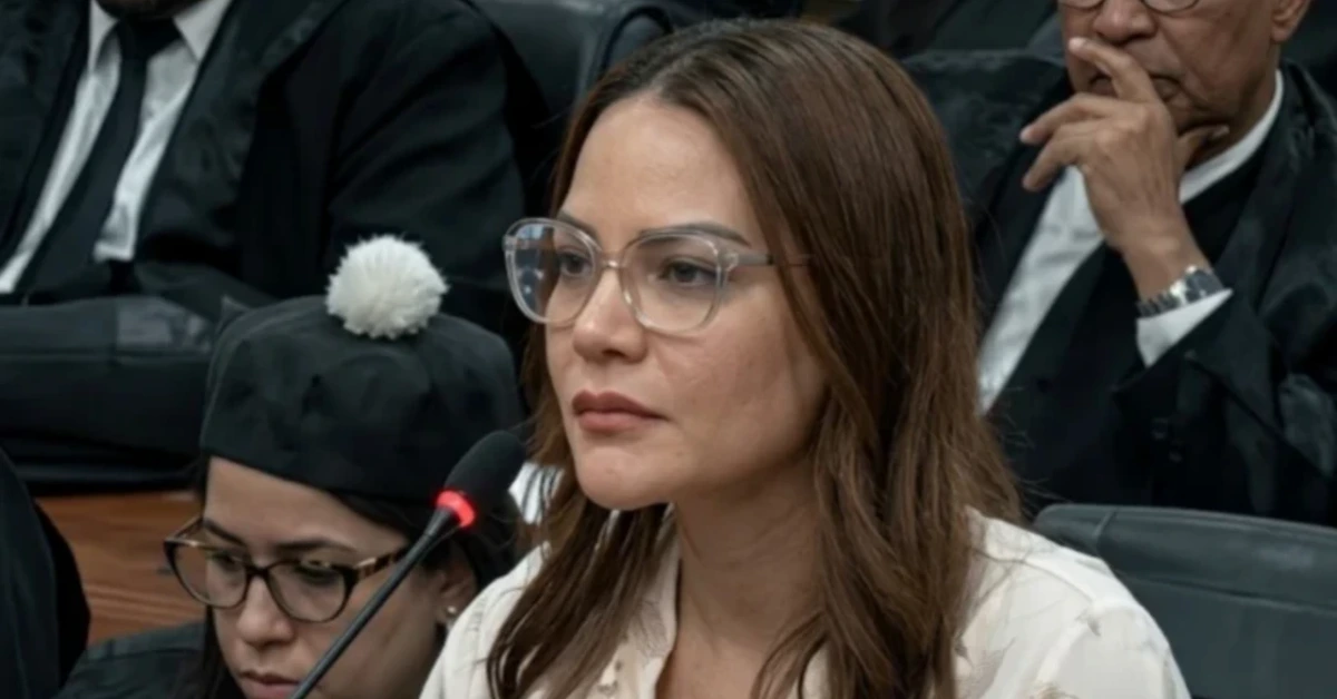 Woman with brown hair and clear glasses speaks into a microphone in a formal hearing, others in robes in the background.