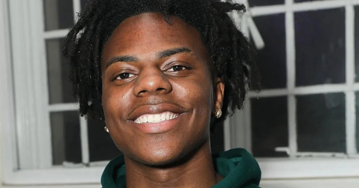 Close-up portrait of a smiling person with short dreadlocks wearing a green hoodie, standing in front of a windowed background