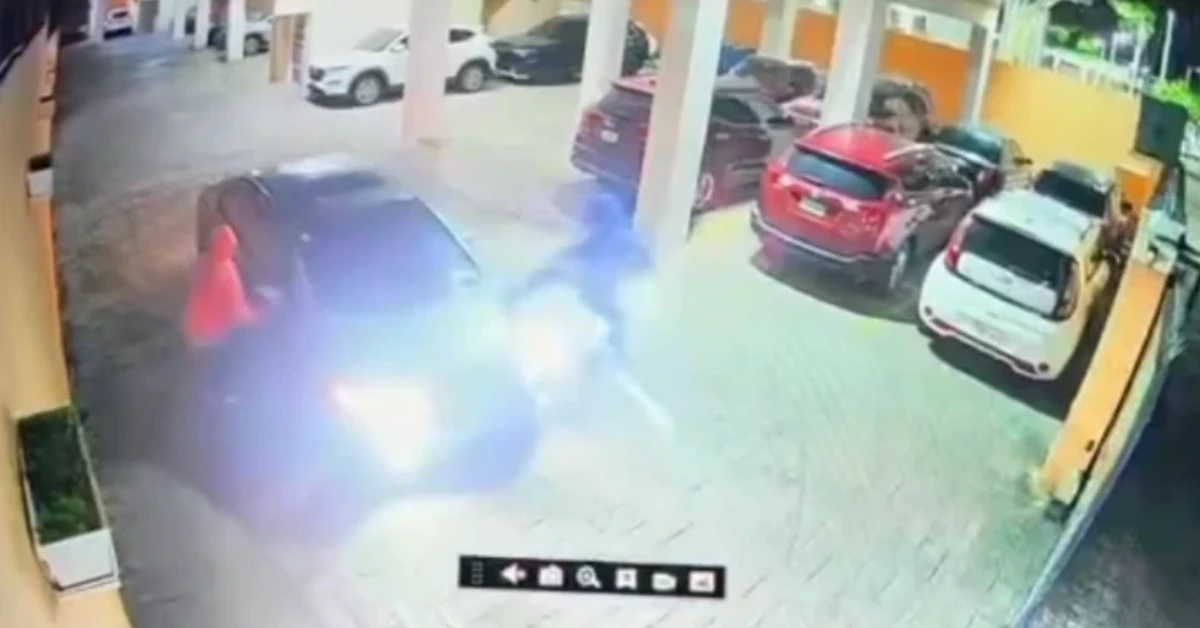 Overhead security camera view of a bright flash and smoke near a dark blue car in a parking area, with several parked cars and yellow columns nearby.