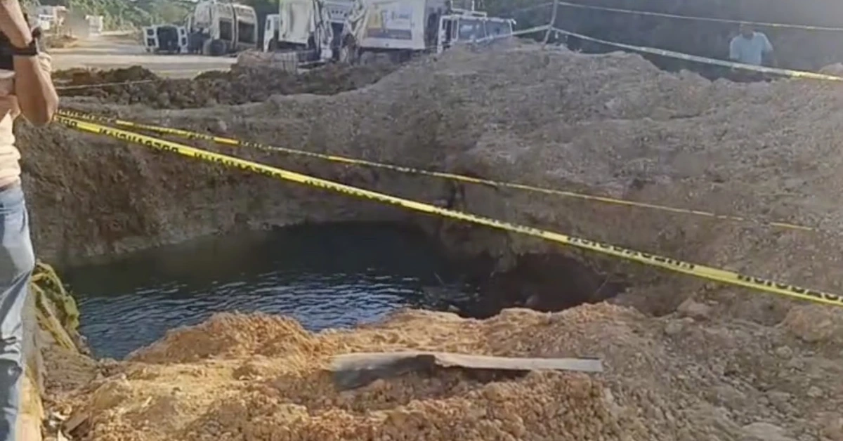 Open muddy sinkhole filled with water at a construction site, surrounded by yellow caution tape and dirt piles; workers and trucks in the background.