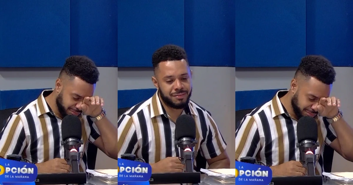 Man in a striped shirt wipes a tear while seated at a microphone on a blue-media studio set.