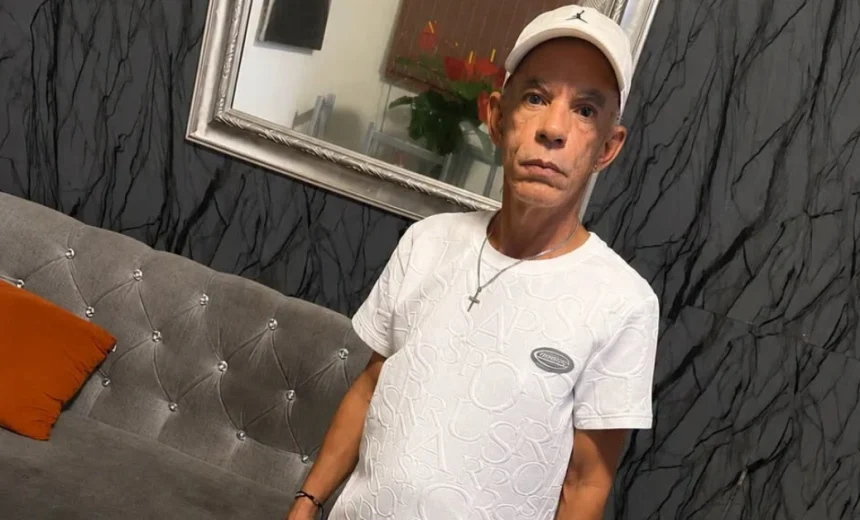 Man in a white cap and textured white T‑shirt with a cross necklace stands beside a tufted gray sofa with an orange pillow in a stylish room.