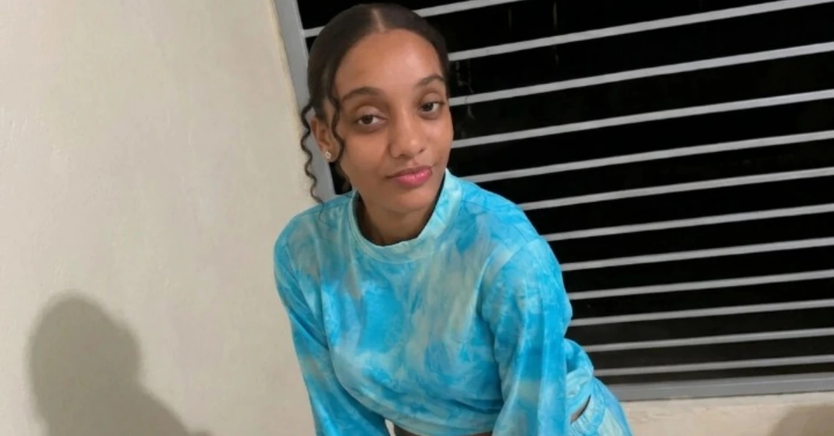 Young woman with tied-back hair wearing a blue tie-dye long-sleeve shirt, leaning forward indoors beside a striped window blind? background.