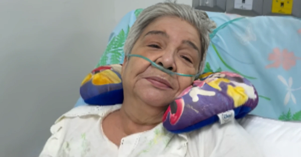 Older woman resting in a hospital bed with a nasal oxygen tube and colorful pillows behind her head.